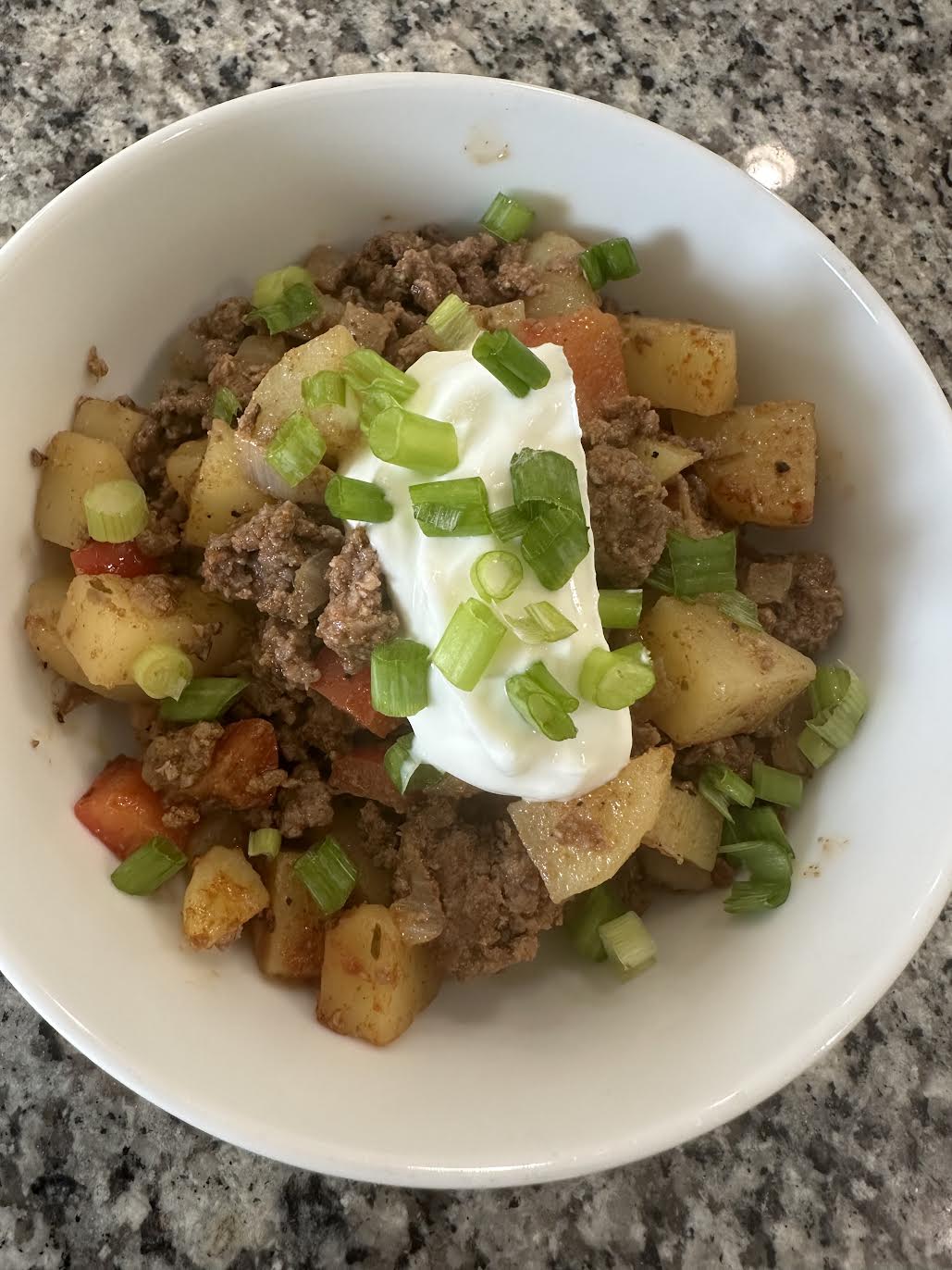 A bowl of ground beef, potatoes, and peppers topped with sour cream and chopped green onions on a granite countertop.