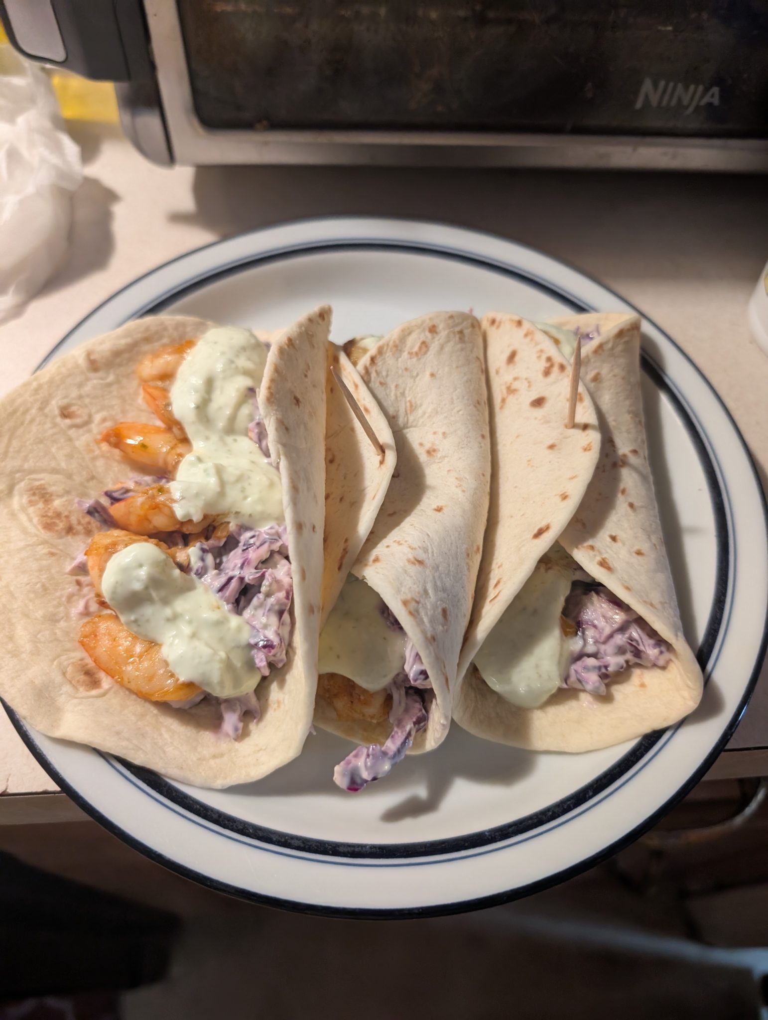 Three soft tacos filled with shrimp, purple cabbage slaw, and a creamy sauce are served on a white plate with a blue rim. The background shows a kitchen counter with a toaster oven.