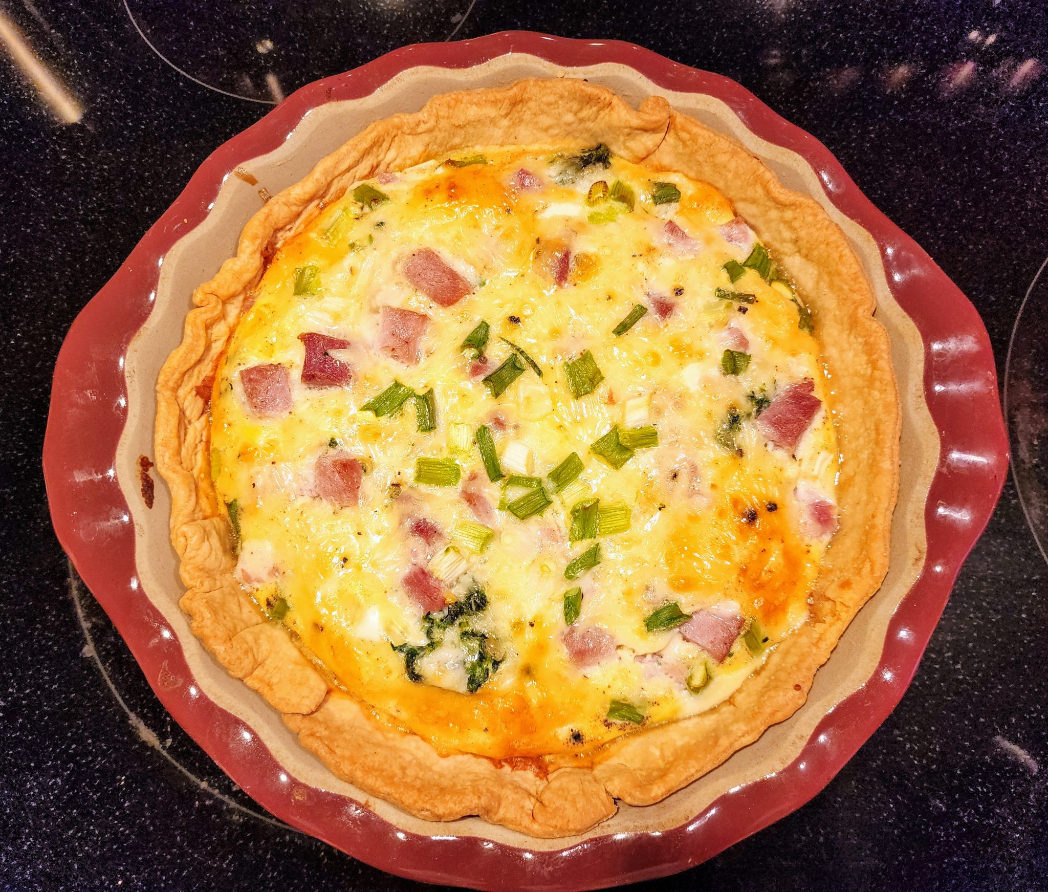 A freshly baked quiche in a red ceramic pie dish, filled with diced ham, green onions, cheese, and eggs, sitting on a black stovetop.