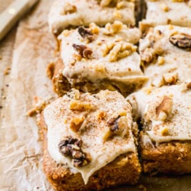 Squares of frosted cake topped with chopped walnuts and a sprinkle of cinnamon are arranged on parchment paper, with one piece slightly pulled away from the group. A knife is visible in the background.