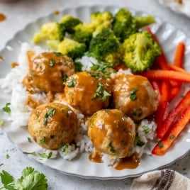 A plate with rice topped with Asian chicken meatballs, surrounded by steamed broccoli and sliced red bell peppers, garnished with chopped herbs. A beige napkin is placed beside the plate, and a small bowl of sauce is in the background.