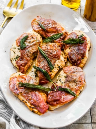 Cooked chicken breasts topped with crispy prosciutto and fresh sage leaves on a white serving platter.