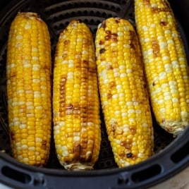 Four ears of roasted corn on the cob with char marks are arranged inside an air fryer basket.