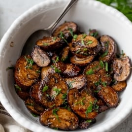 A white bowl filled with sliced air fryer mushrooms garnished with chopped parsley. A spoon rests inside the bowl, and fresh parsley is visible in the background. The surface is light gray, creating a contrast with the dish.