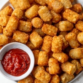 A plate filled with crispy, golden tater tots is shown. A small white bowl of red ketchup is placed on the plate for dipping. The background is a light, neutral color.