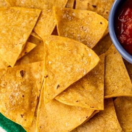 A green plate filled with salted air fryer tortilla chips, arranged in overlapping layers. A small blue bowl in the corner contains red salsa, adding a pop of color to the golden and rustic chips.