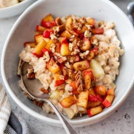 A bowl of apple cinnamon oatmeal topped with diced apples, chopped pecans, and cinnamon, with a spoon resting inside.