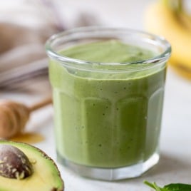 A glass filled with a thick avocado smoothie sits on a light surface, surrounded by a halved avocado, fresh spinach leaves, a honey dipper, and a banana in the background.