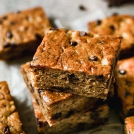 A close-up of several stacked, golden-brown chocolate chip banana bars on a light surface, with chocolate chips visible throughout the soft, moist texture.