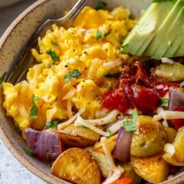 A breakfast bowl of scrambled eggs, roasted potatoes, avocado slices, cheese, salsa, and herbs, with a fork on the side.