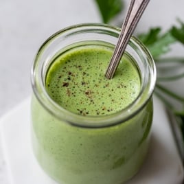 A small glass jar filled with creamy green buttermilk ranch dressing, topped with cracked pepper, and a spoon inside. The jar sits on a hexagonal white surface, with fresh parsley leaves in the background.