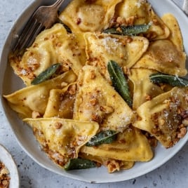 A bowl of butternut squash ravioli with sage brown butter sauce and crushed walnuts, topped with grated cheese. The dish is served on a round plate with a fork on a gray textured surface. Small bowls containing grated cheese and walnuts are nearby.