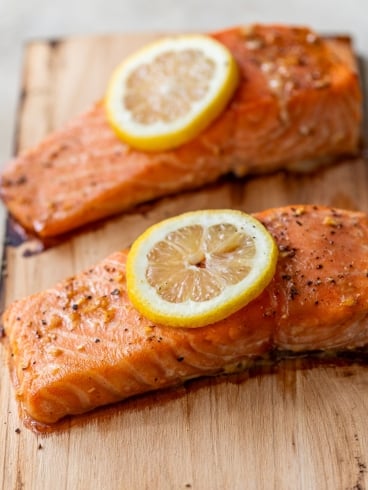 Two cooked salmon fillets on a cedar plank, each topped with a slice of lemon and sprinkled with herbs and pepper.