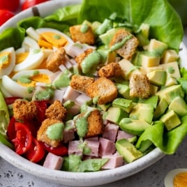 A bowl of chef salad containing lettuce, sliced boiled eggs, cherry tomatoes, cubes of ham and cheese, avocado chunks, croutons, and a drizzle of green dressing. An avocado half and a jar of dressing are in the background on a gray surface.