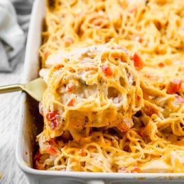A spatula serves cheesy baked chicken spaghetti with diced tomatoes from a white casserole dish.