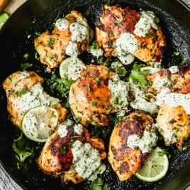 Cilantro lime chicken thighs garnished with fresh cilantro and lime slices, served in a black skillet and topped with a creamy green sauce. The dish sits on a wooden surface with a textured gray cloth underneath.
