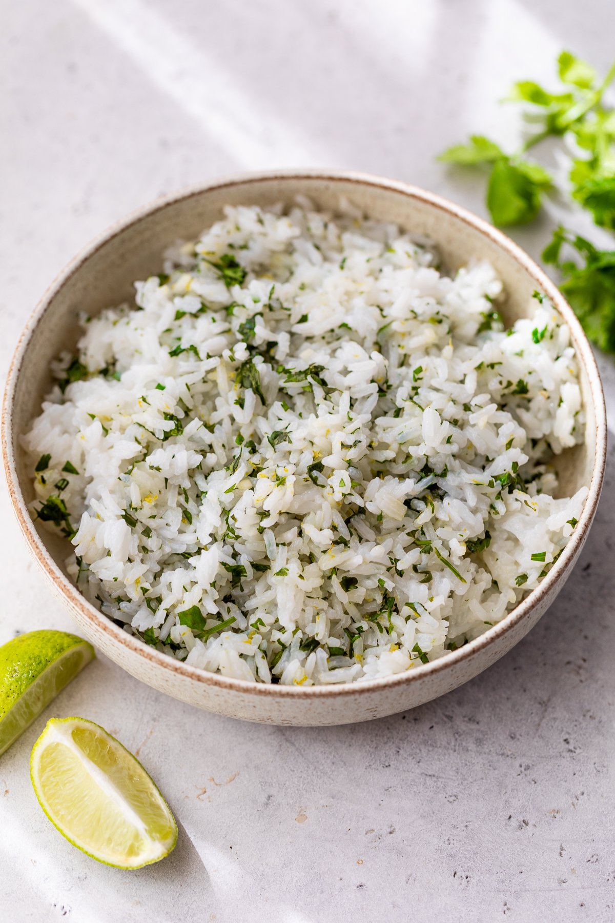 A bowl of fluffy cilantro lime rice, placed on a light-colored surface, with lime wedges and fresh cilantro in the background.