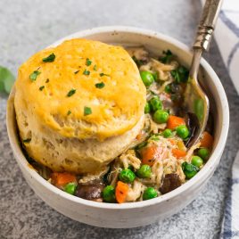 A bowl of chicken pot pie filling with peas and carrots, topped with a golden biscuit and garnished with chopped herbs.