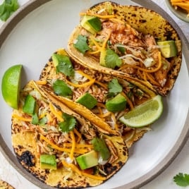 Three crockpot chicken tacos filled with shredded chicken, shredded cheese, avocado chunks, fresh cilantro, and lime wedges on a gray-rimmed plate. Some cilantro and food are scattered on the white surface nearby.