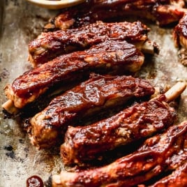 A tray of glazed barbecue crockpot ribs with a shiny, dark red sauce, some ribs stacked and others spread out, next to a small bowl of extra sauce and a basting brush.