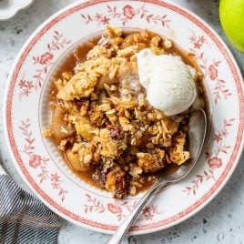 A plate of gluten free apple crisp with a scoop of vanilla ice cream, a spoon, and fresh apples on the side.