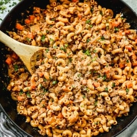 A large black skillet filled with ground beef pasta, topped with grated cheese and chopped parsley. A wooden spoon rests in the pasta, and a striped towel sits nearby.