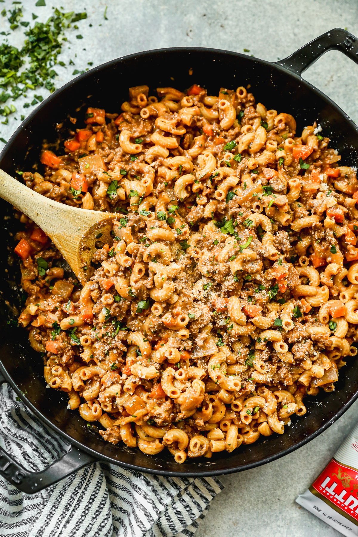 A large black skillet filled with ground beef pasta, topped with grated cheese and chopped parsley. A wooden spoon rests in the pasta, and a striped towel sits nearby.