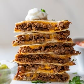A stack of cheese and beef quesadillas cut into wedges, topped with a dollop of sour cream and garnished with cilantro. A bowl of guacamole and some herbs are visible in the foreground.