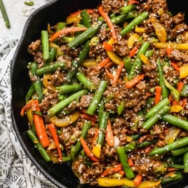 A skillet filled with ground beef stir fry with green beans, red and yellow bell peppers, garnished with sesame seeds and sliced green onions. A patterned cloth is partially visible beneath the skillet.