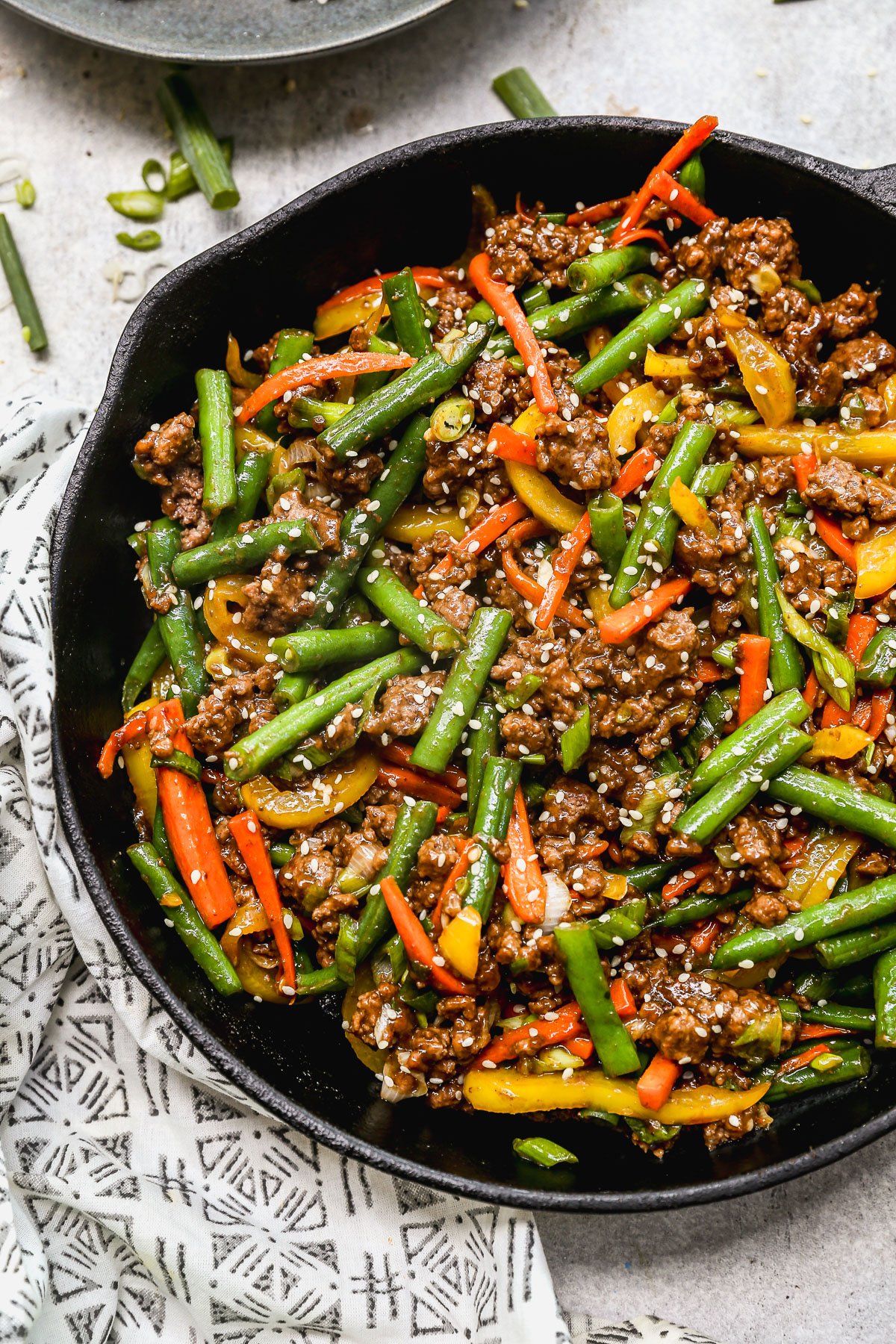 A skillet filled with ground beef stir fry with green beans, red and yellow bell peppers, garnished with sesame seeds and sliced green onions. A patterned cloth is partially visible beneath the skillet.
