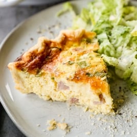 A slice of ham and cheese quiche on a grey plate with a side of green lettuce salad. The quiche has a golden crust and a creamy filling with visible herbs. A striped napkin is partially visible in the corner.