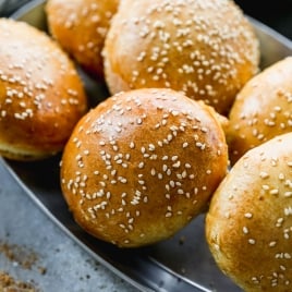 A metal tray filled with freshly baked hamburger buns topped with sesame seeds. The buns are golden-brown, and a sliced tomato is visible in the background.