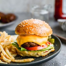 A cheeseburger with lettuce, tomato, and a sesame seed bun on a black plate, accompanied by a side of seasoned fries. A blurred ketchup bottle and two jars are visible in the background on a gray surface.