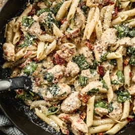 A skillet filled with creamy high protein pasta, chunks of chicken, spinach, and sun-dried tomatoes, topped with grated Parmesan cheese.