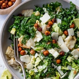 A bowl of kale salad topped with shaved Parmesan cheese, crispy chickpeas, and croutons. A fork rests in the bowl. Lime wedges and a small bowl of dressing are on the side. The background is a light-colored surface.