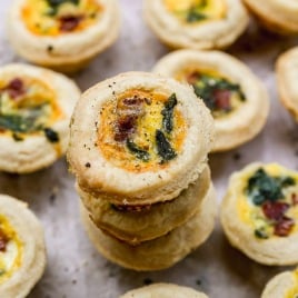 Mini quiche bites with golden crusts, filled with egg, herbs, and bits of bacon, arranged on a light surface.
