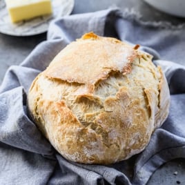 A round loaf of rustic, crusty no-knead bread sits on a gray cloth, with a plate of butter and a butter knife in the background on a gray surface.