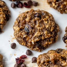 A close-up of oatmeal protein cookies with chocolate chips and dried cranberries on a white surface, with a few chocolate chips and cranberries scattered nearby.