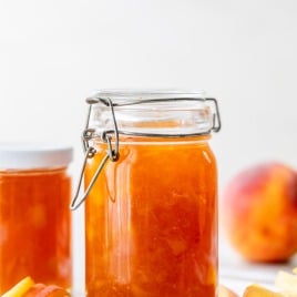 A glass jar of peach jam with sliced peaches in front, and another jar and a peach in the background.
