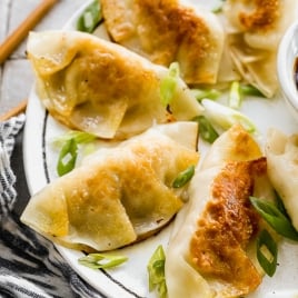 A plate of golden-brown potstickers garnished with sliced green onions. The pork dumplings have crispy edges and are arranged in a semi-circle. A pair of chopsticks is partially visible in the background.