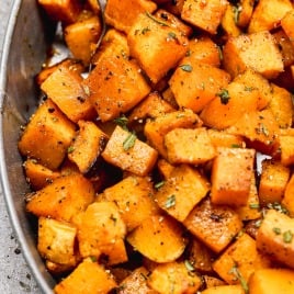 A close-up of roasted butternut squash cubes seasoned with herbs and pepper in an oval dish with a spoon.