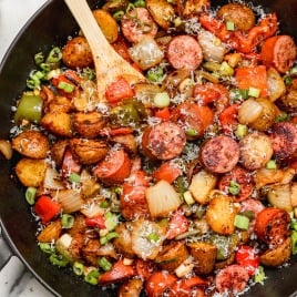 A skillet filled with sautéed sausage slices, diced potatoes, onions, bell peppers, and tomatoes, garnished with chopped green onions and grated cheese, with a wooden spoon resting inside.