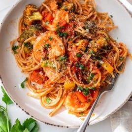 A plate of spicy shrimp pasta with tomato sauce, shrimp, broccoli, bell peppers, and parsley, with a fork on the plate.