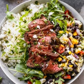 A bowl filled with white rice, topped with slices of grilled steak, arugula, and a mix of black beans, corn, red onion, and peppers. A small bowl of sliced red onions is in the background. The dish is garnished with herbs.