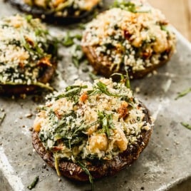 Four stuffed portobello mushrooms topped with breadcrumbs and fresh herbs on a rustic metal tray.
