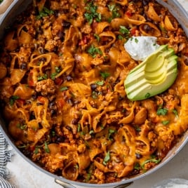 A skillet filled with cheesy taco pasta shells, black beans, and ground meat, topped with sliced avocado, chopped cilantro, and a dollop of sour cream. A striped towel and a bowl of sour cream are nearby.