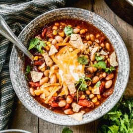 A bowl of taco soup topped with shredded cheese, tortilla chips, cilantro, and sour cream sits on a wooden table.