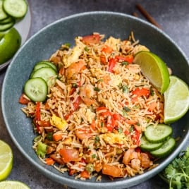 A bowl of Thai fried rice garnished with lime wedges and cucumber slices, surrounded by fresh lime halves and cilantro on a gray surface.