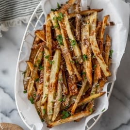 A white basket with parchment paper holds a serving of golden, crispy truffle fries garnished with chopped parsley. The background features a striped cloth, a small bowl of grated cheese, and a glass of oil on a marble surface.