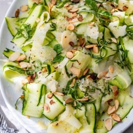 Thinly sliced zucchini ribbons with shaved cheese, toasted almonds, basil, and lemon on a white plate.